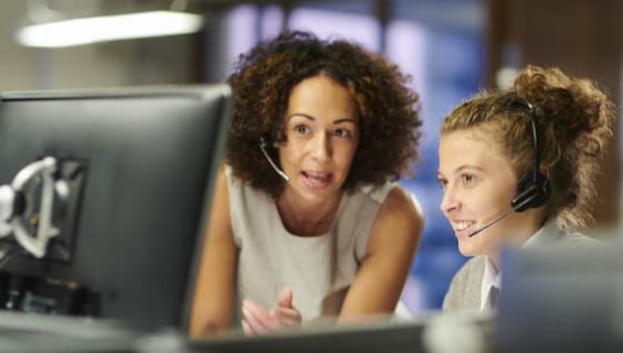 Two women at computer with headphones. One is standing the other sitting.