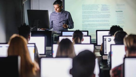 Trainer facing toward us in front of the class with several students sitting at computers facing him