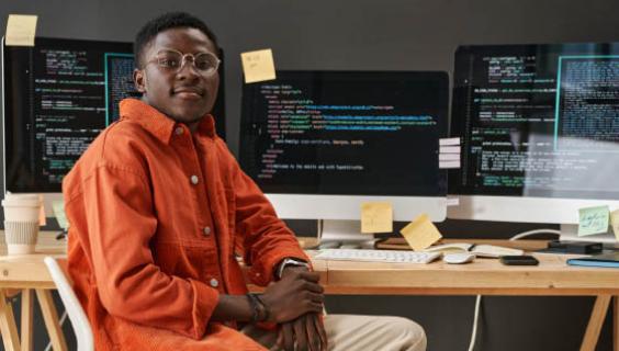 Boy with glasses looks at camera with three computer screens behind him